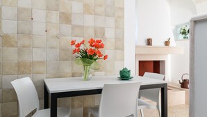 Ein Essbereich mit einer Fliesenwand, einem Tisch mit Stühlen, einer Vase mit Blumen und einem Kamin mit Wasserkocher.