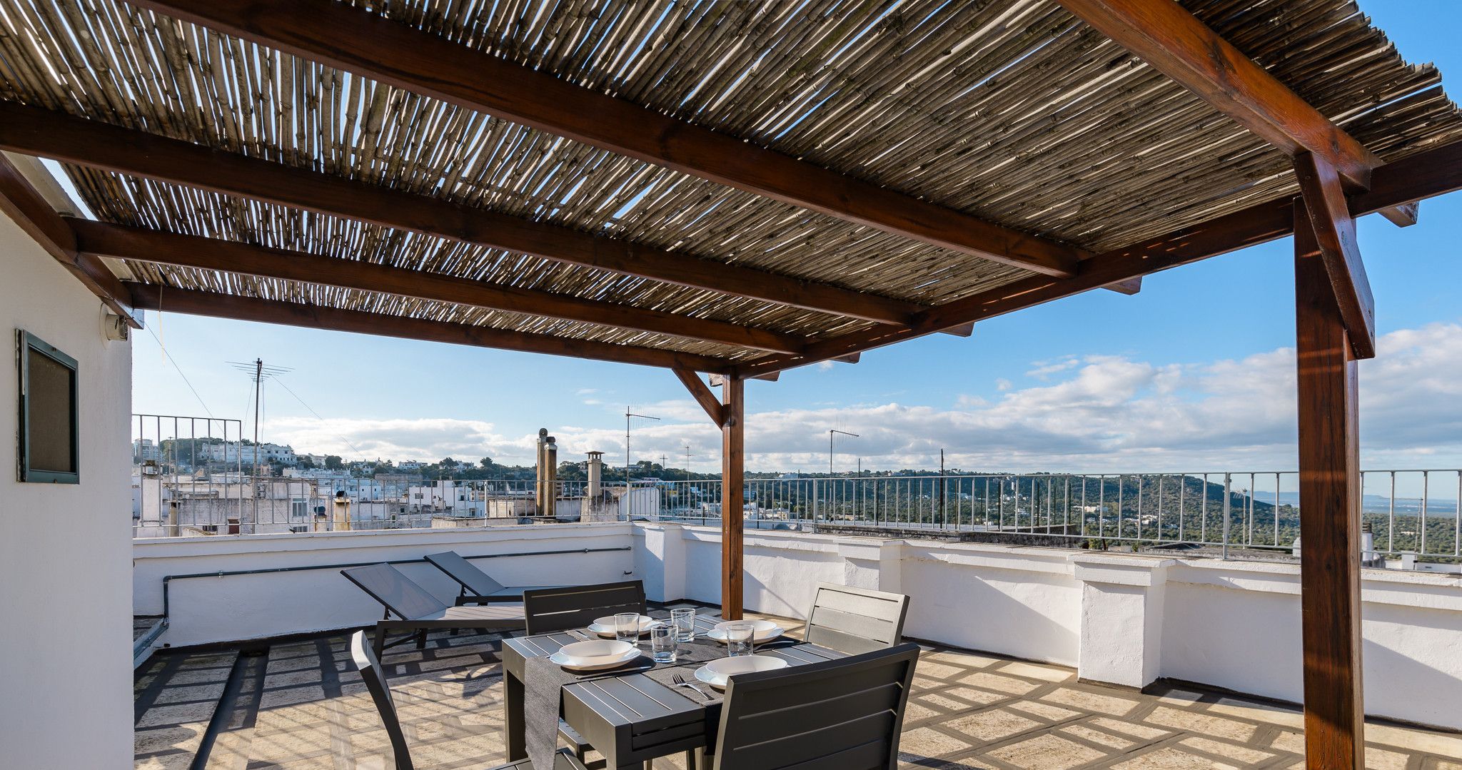 Une terrasse sur le toit, avec une table dressée pour le dîner, une pergola au-dessus et une vue sur la ville en arrière-plan.