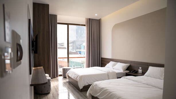 A hotel room with two beds, a large window with curtains, a wooden floor, and a view of buildings outside.