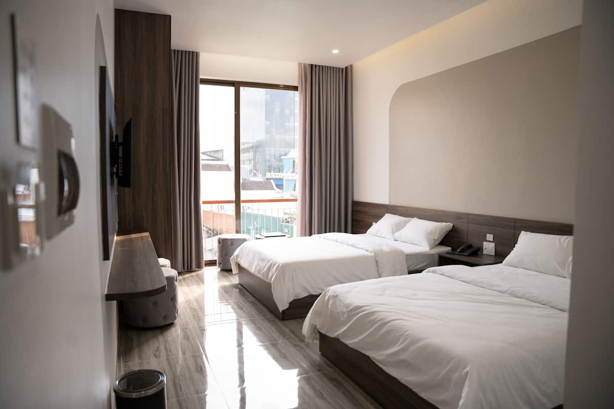 Une chambre d’hôtel avec deux lits, une grande fenêtre avec des rideaux, un plancher en bois et une vue sur les bâtiments environnants.