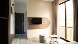 A modern hotel room with a large window, a flat-screen TV mounted on the wall, a round ottoman, and a small round table.