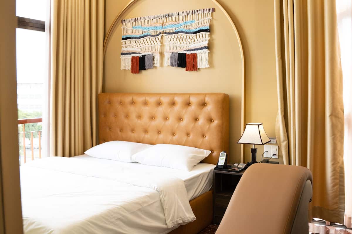 Une chambre d’hôtel avec une tête de lit capitonnée, un lit avec du linge de lit blanc, un fauteuil aux lignes courbes, une table de chevet avec une lampe et une décoration murale en macramé.