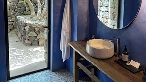 Une salle de bain avec un mur en pierre, un miroir rond, un meuble-lavabo en bois et une vue sur un mur en pierre et un arbre à l’extérieur.