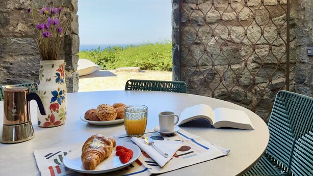 Une table avec des croissants, des fraises, un verre de jus d’orange et un livre, le tout placé devant un mur de pierre avec vue sur la mer.