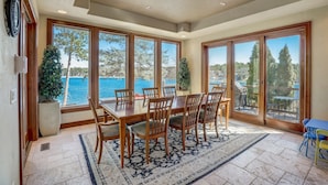 A dining area with a large table and chairs, large windows with a view of the sea, and a sliding glass door leading to an outdoor patio. - Luxury Lakefront! Hot Tub & Arcades! Sleep 24! (Wautoma)