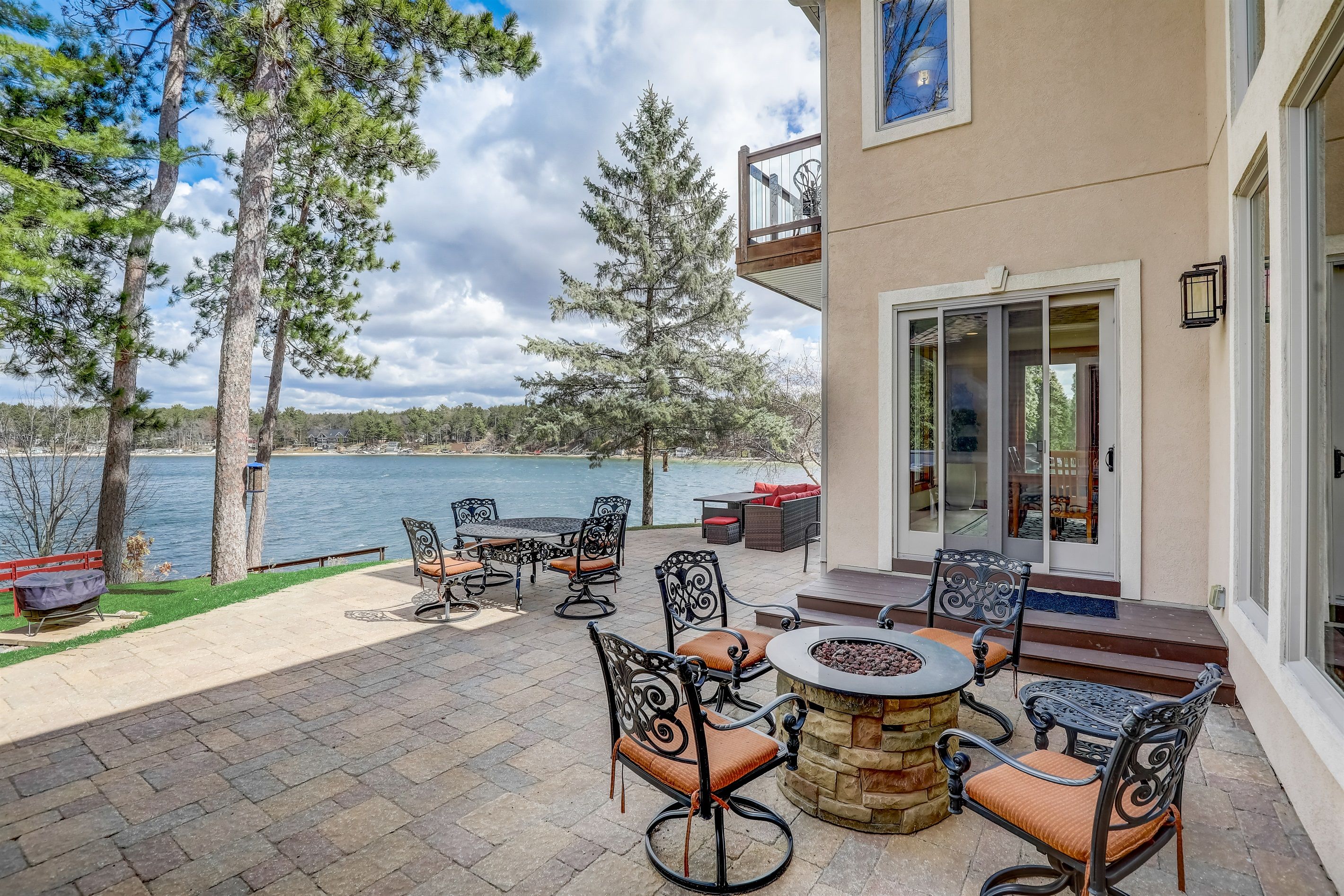 An outdoor patio with a stone fire table, metal chairs, and a view of a lake.