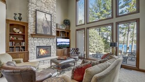 A cozy living room with a stone fireplace, leather furniture, and large windows overlooking a scenic view. - Luxury Lakefront! Hot Tub & Arcades! Sleep 24! (Wautoma)