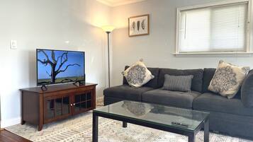 A living room with a flat-screen TV on a wooden cabinet, a glass coffee table, a grey sofa with patterned cushions, and a window with blinds.