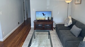 Un salon avec un téléviseur à écran plat posé sur un meuble en bois, un canapé de couleur foncée agrémenté de coussins à motifs, une table basse en verre et un lampadaire.