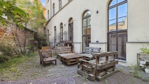 A courtyard with wooden furniture and a patterned rug, surrounded by a brick wall and a building with large windows. - Cityloft Gold - im Zentrum. Inkl. Parken, Netflix (Leipzig)