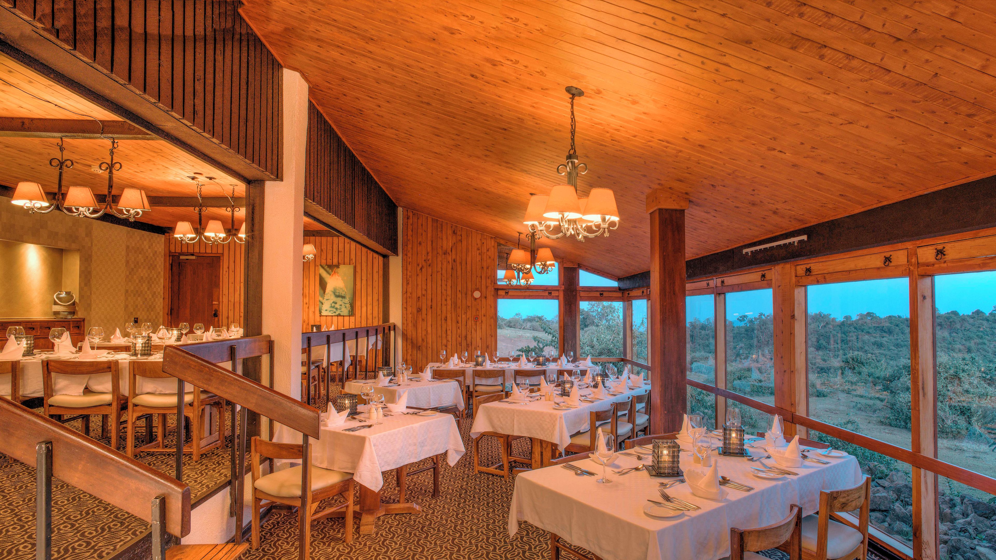 A restaurant with multiple dining tables set for guests, wooden interior, and large windows offering a view of the outdoors.