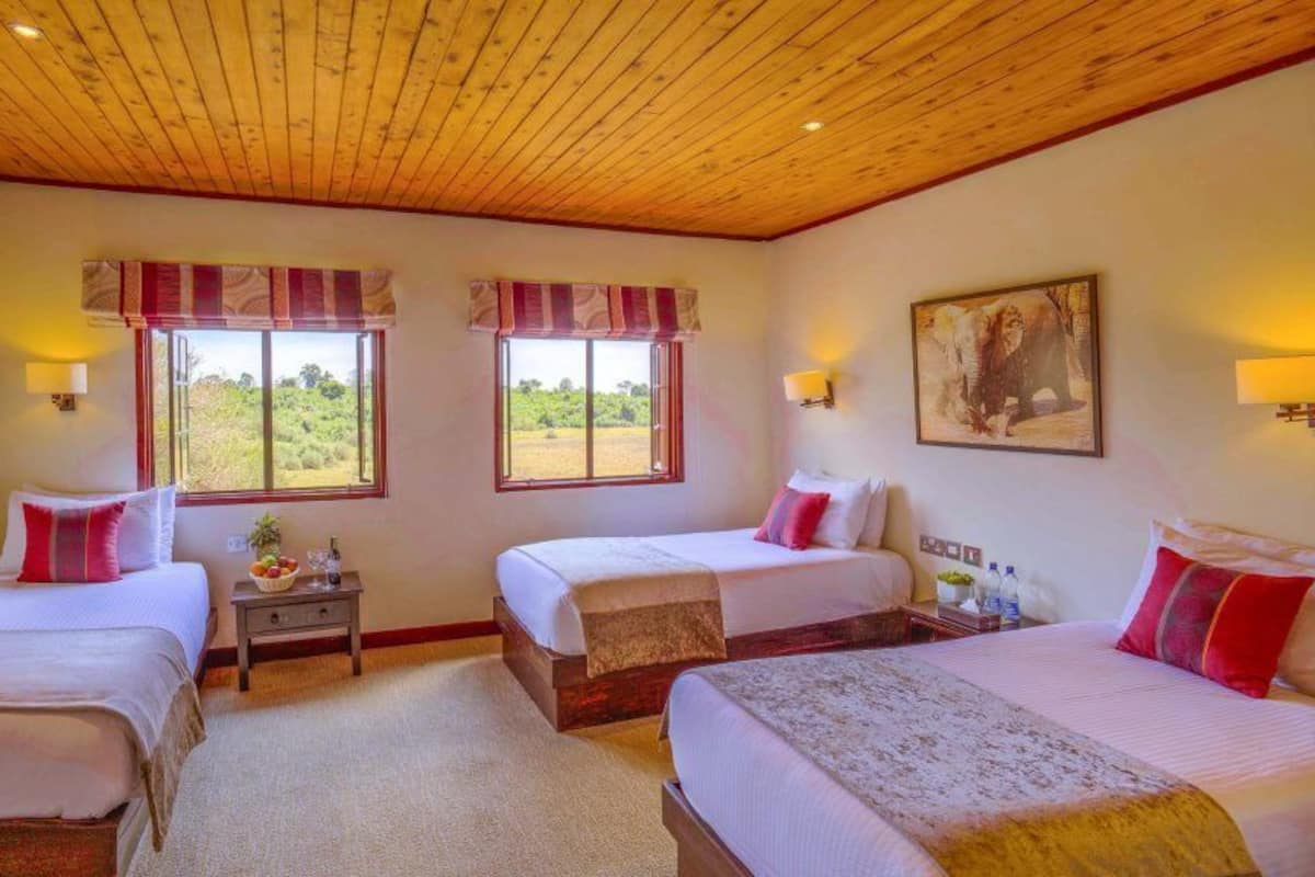 A room with three beds, wooden ceiling, large windows with curtains, wall-mounted lamps, and a painting.