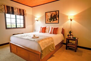 A hotel room with a large bed, two bedside lamps, a window with curtains, a painting on the wall, and a small table with a fruit bowl.