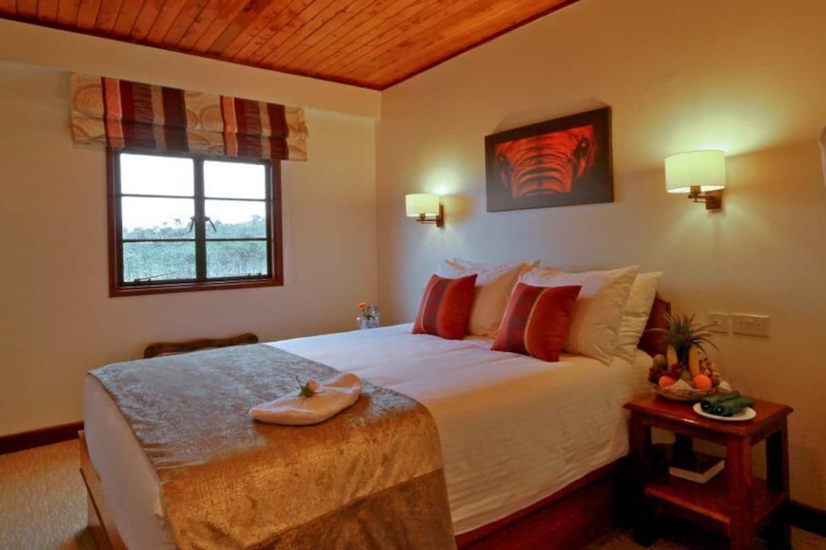 A bedroom with a large bed, two bedside lamps, a window with curtains, a painting on the wall, and a nightstand with a fruit bowl.