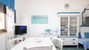 A room with a dining table, chairs, a television, a cabinet with glass doors, a bed, and a wall-mounted decorative item.