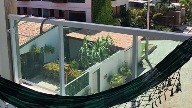 A balcony with a hammock, overlooking a courtyard with plants and a building with balconies. - Pousada Porto dos Reis (Ipojuca)