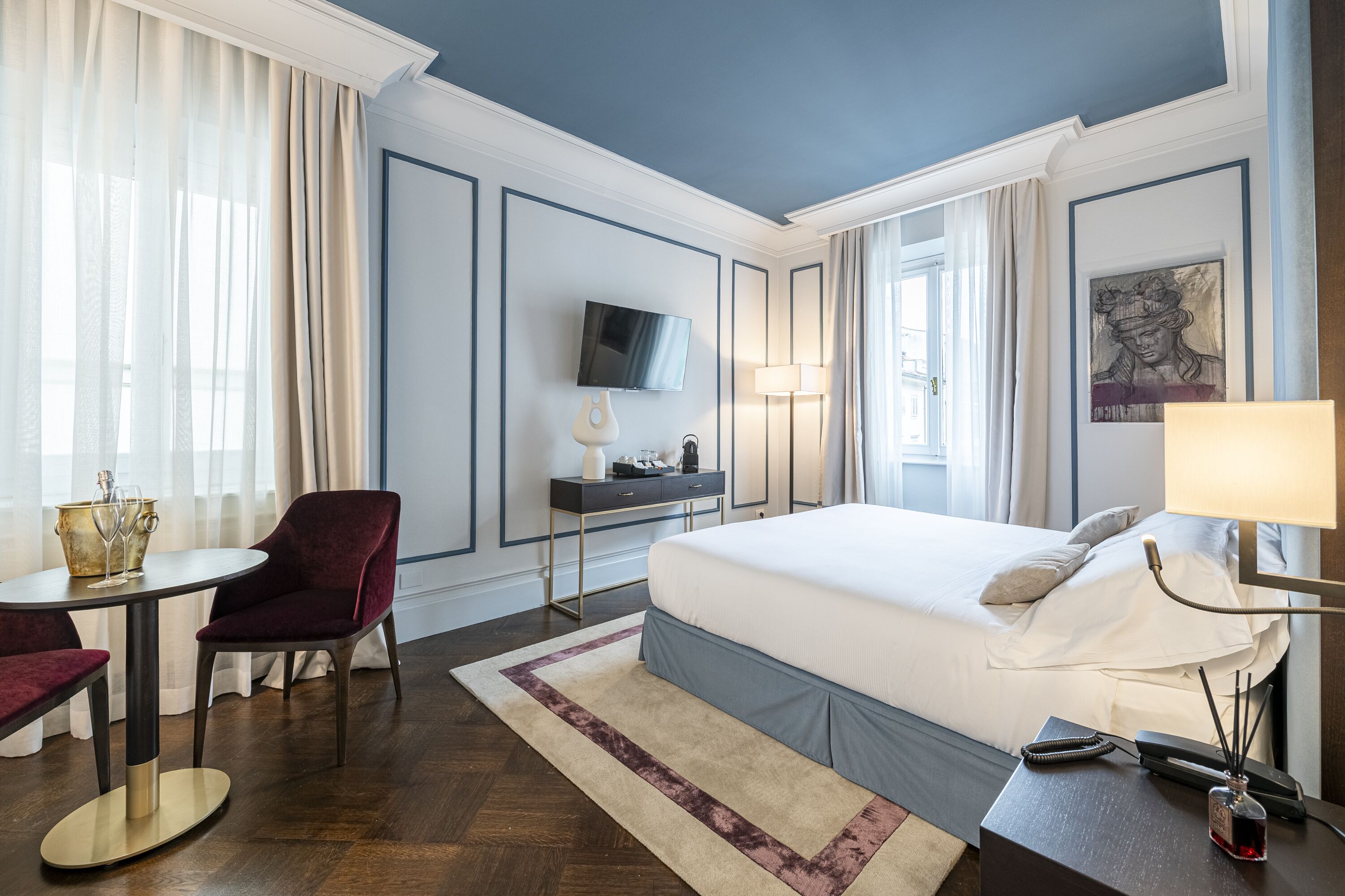 A hotel room with a bed, a desk, a chair, a TV, and a painting on the wall.