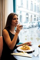 Una persona seduta a un tavolo con un piatto di cibo e un bicchiere, con vista su edifici e auto parcheggiate.
