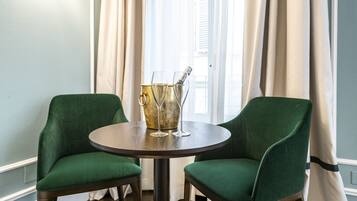 Quarto com mesa redonda, duas cadeiras verdes estofadas, um objeto decorativo dourado e uma janela com cortinas.
