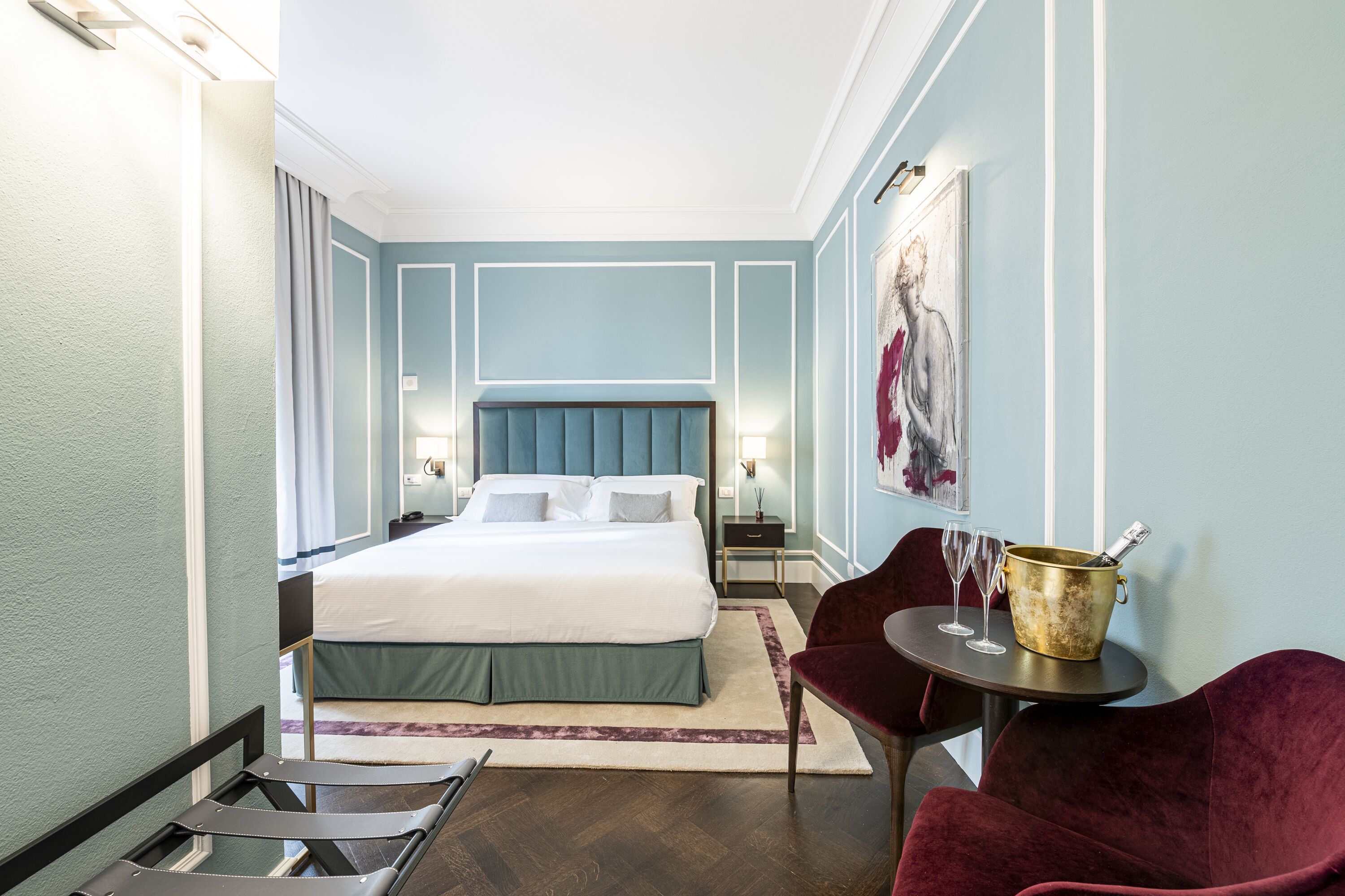 Habitación de hotel con cama, mesita de noche, silla, mesa con un cubo y un cuadro en la pared.