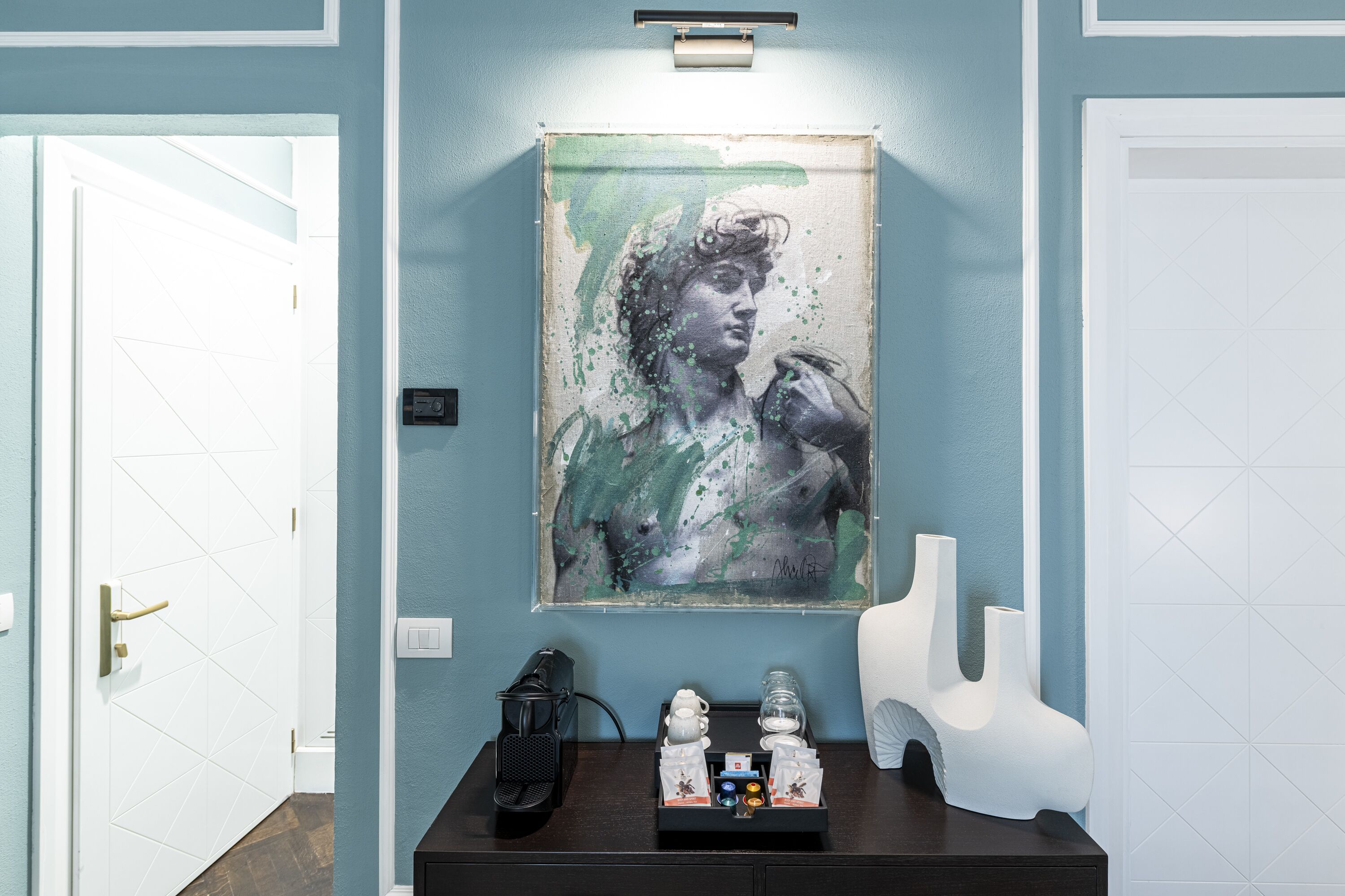 A hallway with a blue wall, a framed artwork, a coffee station, and a white door.