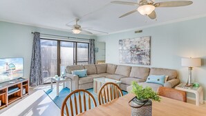 Un salón amplio con un sofá modular grande, una mesa de comedor con sillas, un ventilador de techo y un ventanal con cortinas.