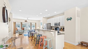 A modern kitchen with white cabinets, a stainless steel refrigerator, and a microwave. There are colorful stools and a wooden dining table with a woven placemat.