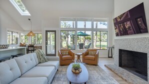 A spacious living room with a sofa, two armchairs, a coffee table with plants, a fireplace, and large windows offering a view of the outdoors.