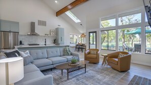 A spacious living area with a sofa, armchairs, and a coffee table. A kitchen with modern appliances and a dining area with a table and chairs are visible. Large windows provide ample natural light and views of the outdoors.