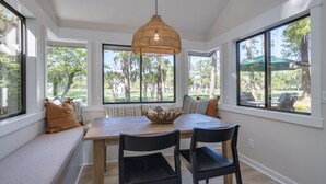 A dining area with a large table, two black chairs, a woven pendant light, and a view of a park through large windows.