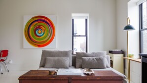 A bedroom with a wooden chest, a bed with grey pillows, a colorful abstract painting, a window, and a floor lamp. - Stylish, spacious suite in Wellington- steps to beach, dining, brewery and shops (Prince Edward)