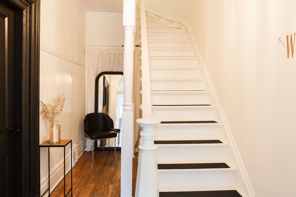 Un escalier avec des marches blanches et des contremarches noires, une chaise noire et un vase contenant des fleurs séchées, posé sur une petite table.