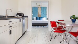 A modern kitchen with white cabinets, a stainless steel sink, and a coffee machine. A dining area with a white table and red chairs is visible. A blue door with shutters leads to a bedroom with a bed and a blue headboard. - Stylish, spacious suite in Wellington- steps to beach, dining, brewery and shops (Prince Edward)