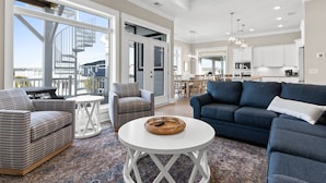 A spacious living room with a blue sofa, striped armchair, and a white coffee table. A large window offers a view of a balcony and outside buildings. - Oyster Perpetual, 5 bedroom 5 bathroom home that ensures your stay is timeless. (Ocean Isle Beach)