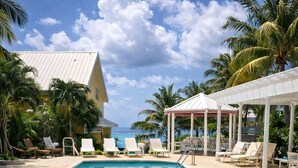 A swimming pool with lounge chairs and a covered seating area, surrounded by palm trees and a building with a sloped roof.