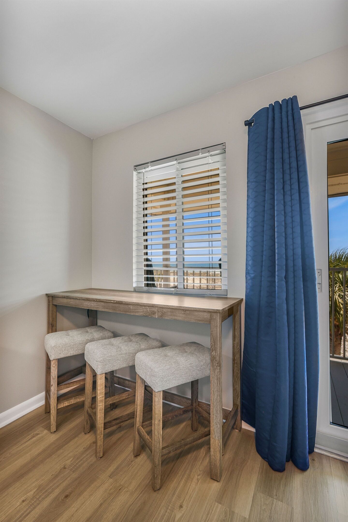 A room with a wooden table, two grey cushioned stools, a window with blinds, and a blue curtain.