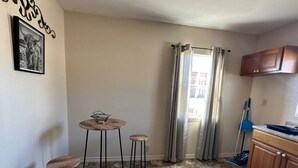 Una cocina pequeña con zona de comedor, que incluye una mesa y taburetes, una ventana con cortinas, y una escoba apoyada en la pared.
