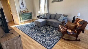 Una sala de estar con un sofá modular, un tapete estampado, una mesa de centro de madera, un sillón mecedor y un aparador verde.