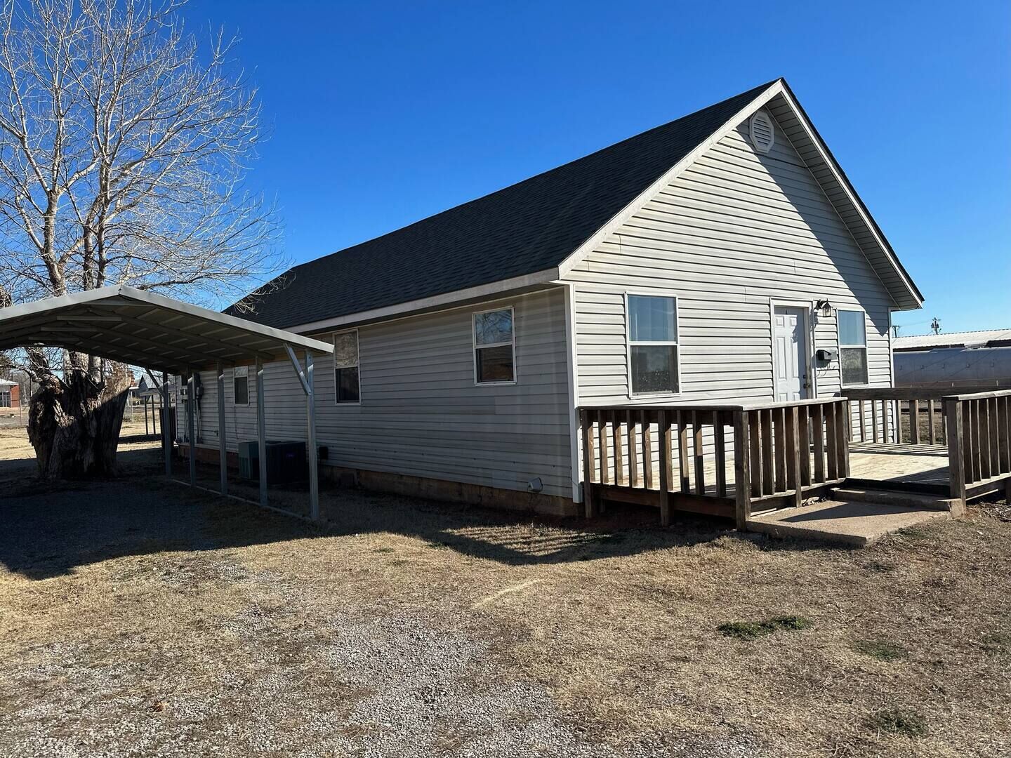 A single-story house with a covered patio, a fenced deck, and a large tree in the background.