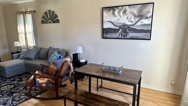 Una sala de estar con un sofá modular, una mecedora, una mesa de comedor con bancos, un ventilador de pared y un cuadro enmarcado de un toro.