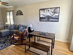 A living room with a sectional sofa, a rocking chair, a dining table with benches, a wall-mounted fan, and a framed picture of a bull.