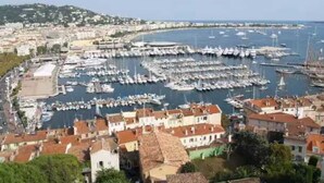Une ville côtière dotée d’un port de plaisance rempli de bateaux, entourée de bâtiments traditionnels aux toits recouverts de tuiles rouges.