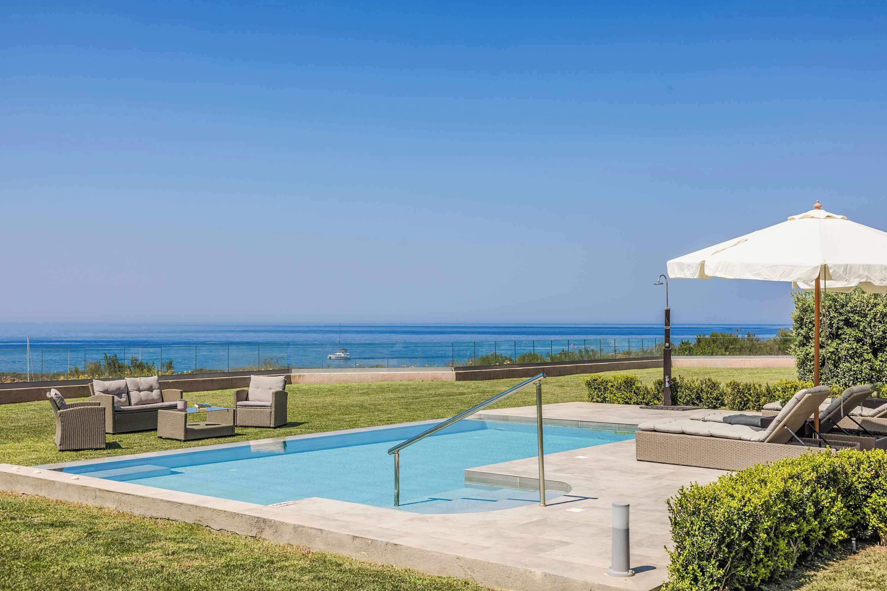 A swimming pool with a view of the ocean, outdoor furniture, and a white umbrella.
