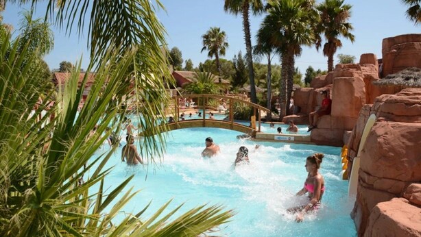 Zona de piscina con gente nadando y relajándose, rodeada de palmeras y formaciones rocosas.