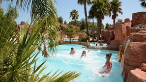 Zona de piscina con gente nadando y relajándose, rodeada de palmeras y formaciones rocosas.