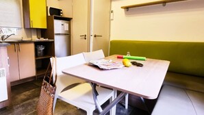A compact kitchen with a microwave, a refrigerator, and a sink. There is a dining table with a magazine and a bottle. A green chair is visible.