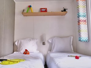 Quarto de hotel com duas camas, um armário com brinquedos, uma luminária na parede e uma cortina estampada.