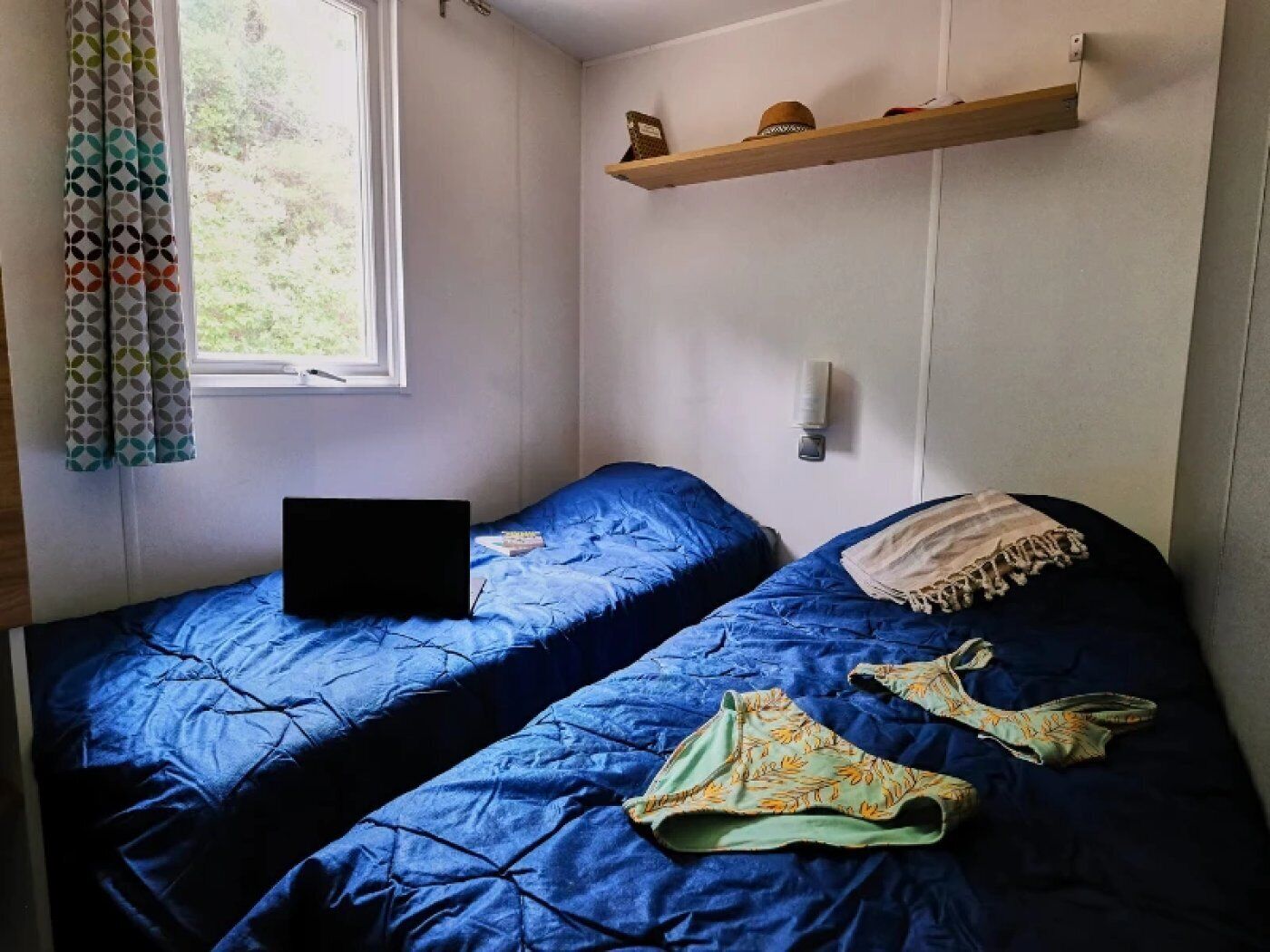 Une chambre avec deux lits, une fenêtre, un ordinateur portable posé sur l’un des lits et une étagère avec divers objets.