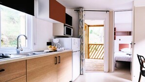 A compact kitchen with a microwave, toaster, and sink. A bed is visible through an open door, and a chair is in the foreground. - ZEcamping 4 étoiles piscine animaux acceptés mobil-home TV LV clim (Orgon)