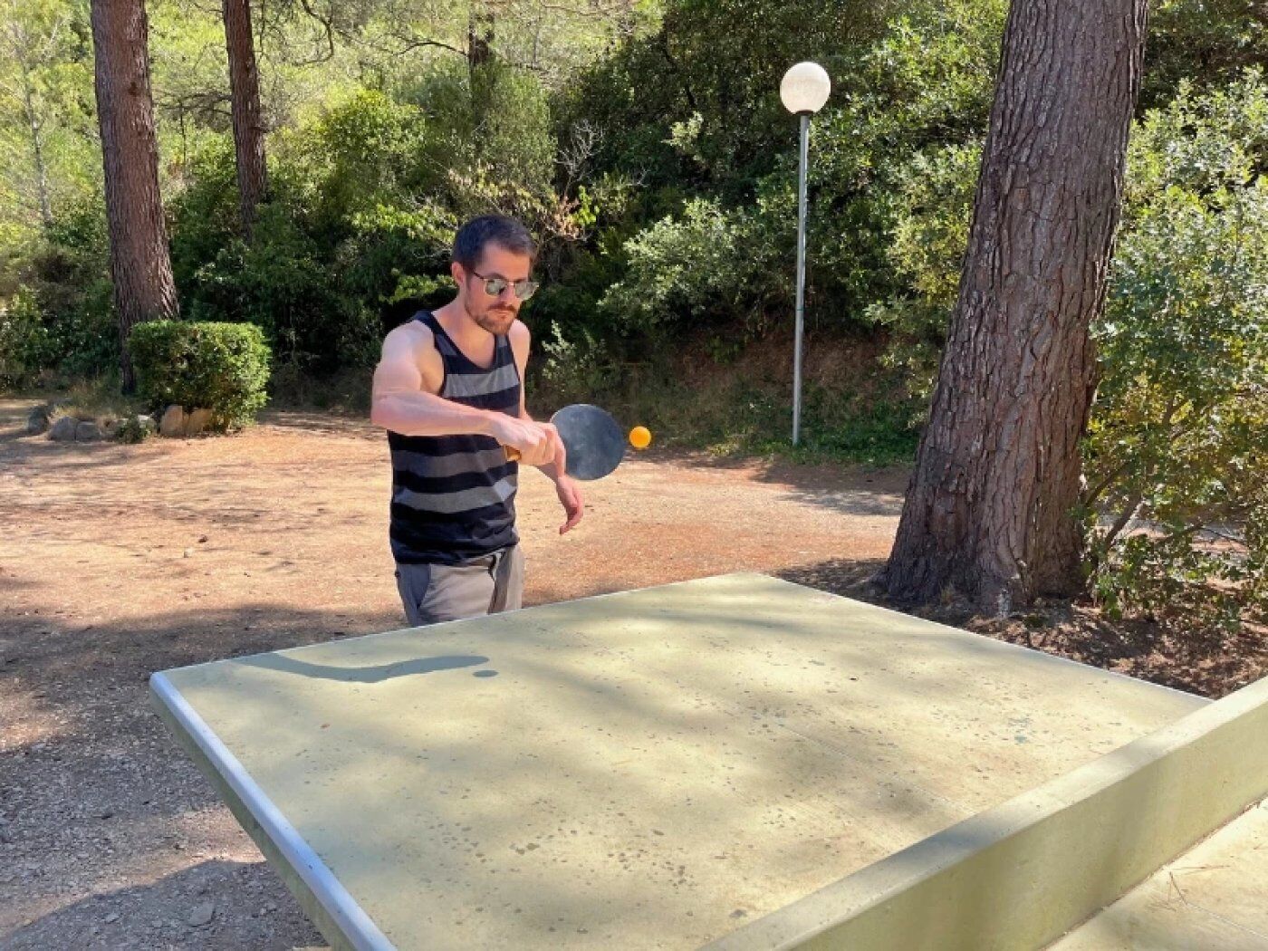 Un homme joue au ping-pong à l’extérieur, sur un terrain en terre, avec des arbres en arrière-plan.
